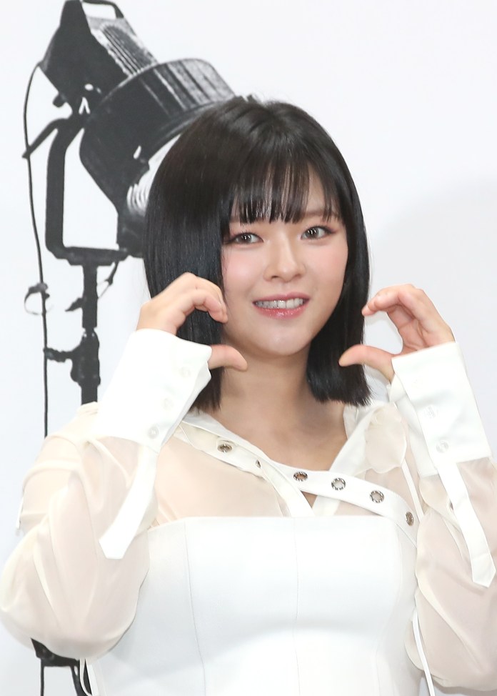 A person with black hair wearing a white outfit makes heart shapes with their hands, smiling at the camera. A studio light silhouette is visible in the background.