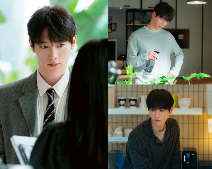 Seo Kang-joon with dark hair wearing a suit and tie is engaged in a serious conversation with someone off-camera, set against a backdrop of lush green plants.