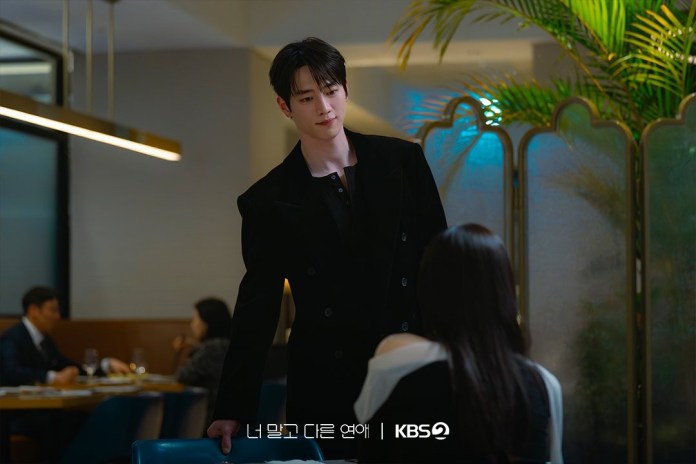 Seo Kang-joon in a stylish black jacket leans slightly over a table, looking at a woman seated in a restaurant. Soft lighting and indoor plants create a cozy atmosphere.