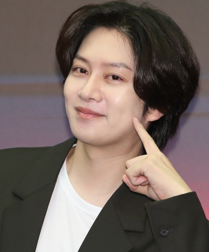 Kim Heechul with dark, wavy hair is smiling and pointing to their cheek, wearing a black blazer over a white shirt.