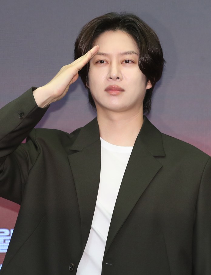 Kim Heechul in a dark blazer and white shirt, performing a salute at an event.
