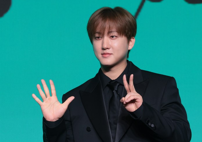 Changbin in a black suit stands against a green background, holding up one hand with five fingers extended and the other hand making a peace sign.