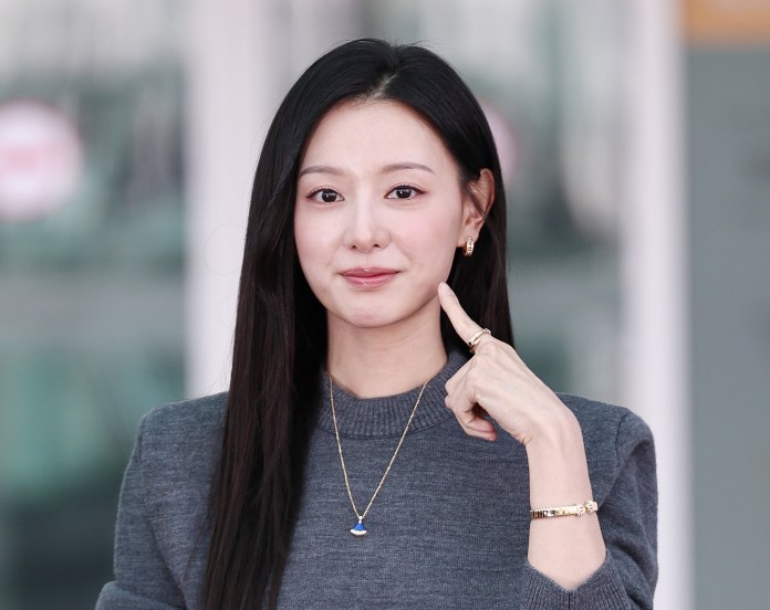 Kim Ji-won with long black hair wearing a gray sweater and gold jewelry, smiling and pointing to her chin.