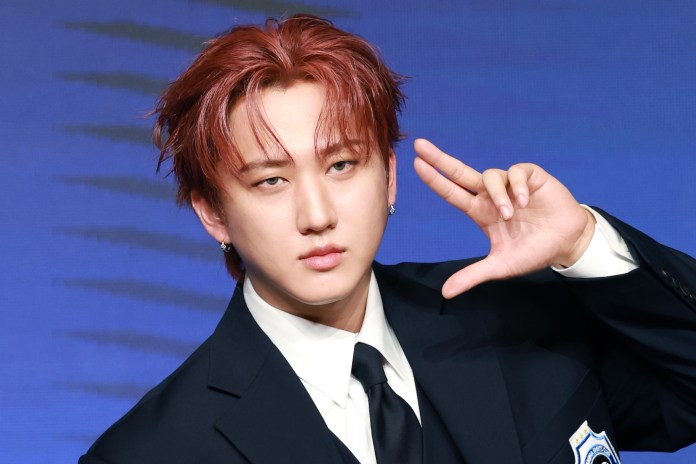 Changbin with reddish hair and earrings poses confidently, wearing a black suit and tie, with a playful hand gesture in front of a blue background.
