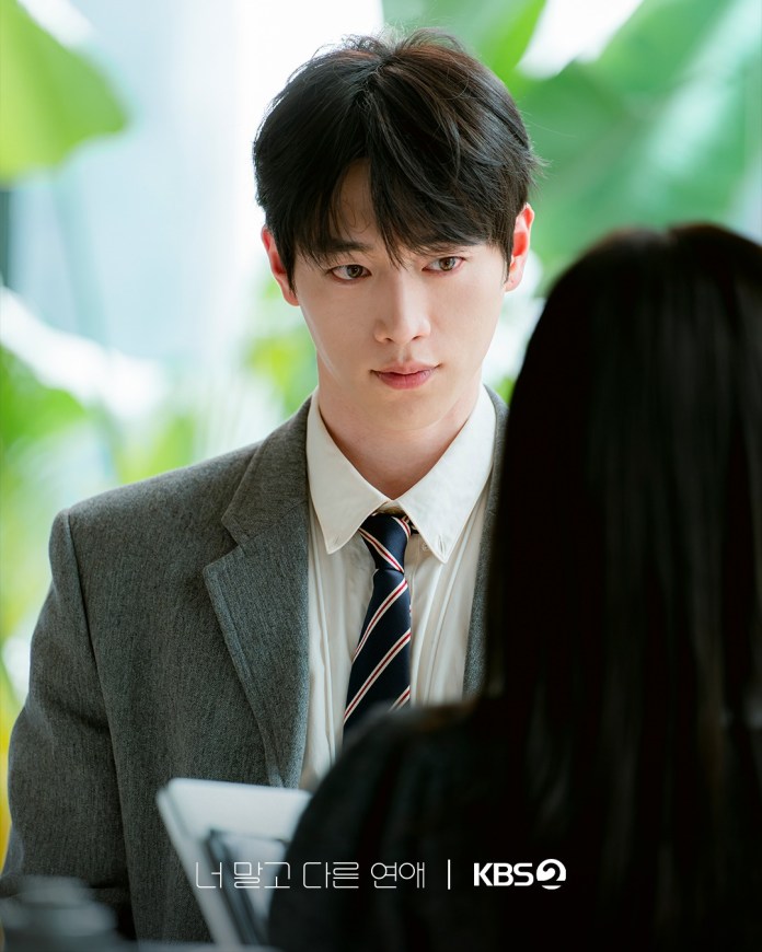 Seo Kang-joon in a suit with short dark hair is looking intently at a woman, whose face is out of view. The background features lush green plants, suggesting a modern office setting.