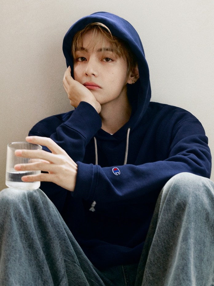BTS V in a navy hoodie sits casually, resting their chin on their hand, holding a glass of water.