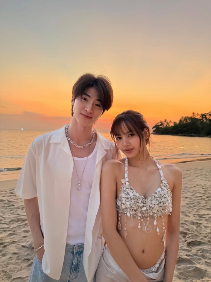 Lisa and Thai Actor Blue Pongtiwat poses for a photo on the beach at sunset, with a colorful sky in the background. The man is wearing a white shirt and denim jeans, while the woman is dressed in a sparkling, intricately designed top and a revealing outfit.
