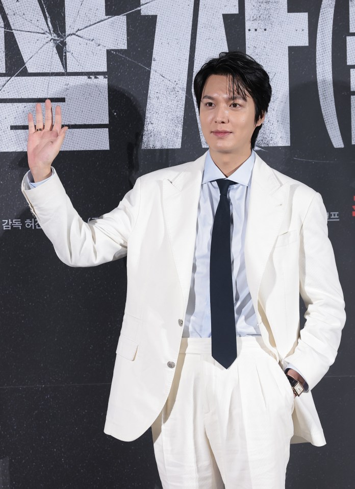 Lee Min-ho in a white suit and blue shirt is waving at the camera, standing in front of a dark background with text graphics.