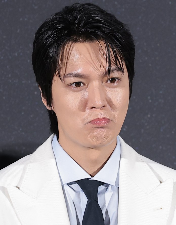 Lee Min-ho with dark hair making a pouting expression, wearing a white suit and a light blue shirt with a dark tie.