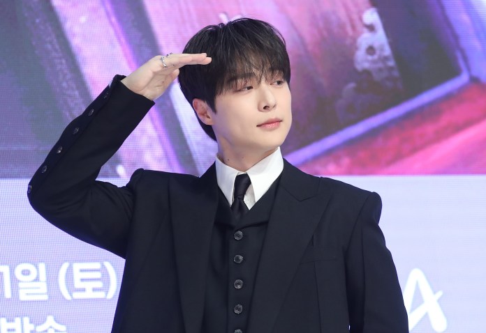 Bae In-hyuk in a formal black suit saluting with a confident expression during a press event.