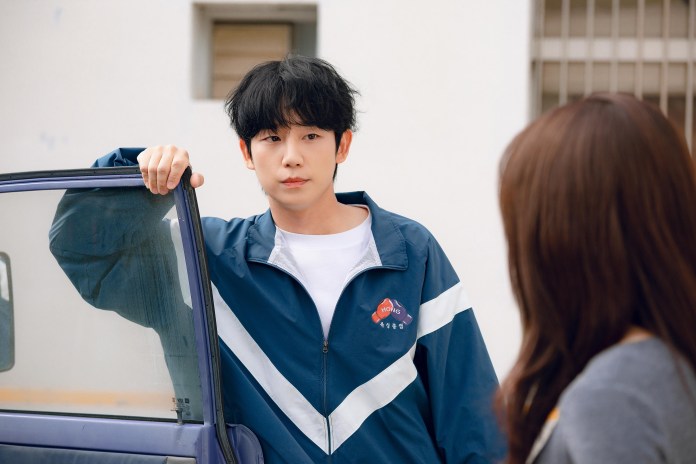 A young man with black hair wearing a blue jacket leans against a car, looking thoughtfully at a woman across from him.