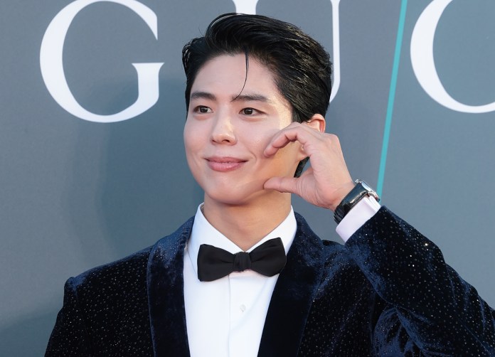 Park Bogum wearing a black velvet tuxedo with a bow tie, smiling and making a heart shape gesture with his hand against a backdrop featuring the word 'Gucci'.