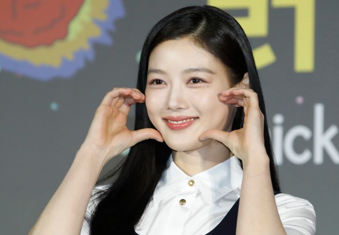 Kim Yoo-jung with long black hair is making a playful gesture by framing her face with her hands, set against a colorful background.