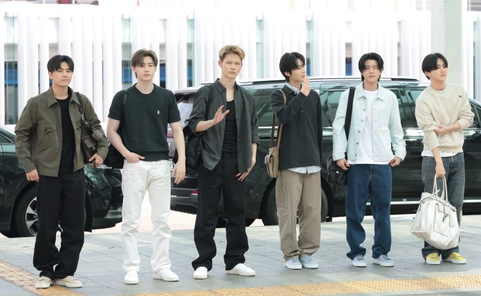 ENHYPEN members standing together outdoors, dressed in casual fashion. They are posing in front of a black vehicle, with a modern building in the background.