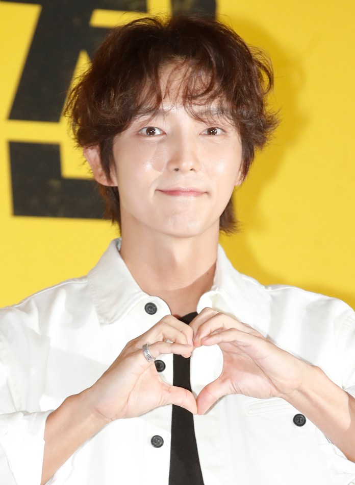 Lee Joon-gi with wavy hair making a heart shape with his hands against a bright yellow background.