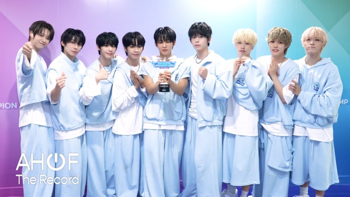 AHOF in matching light blue outfits, standing together and smiling, with one member holding a trophy in the center.