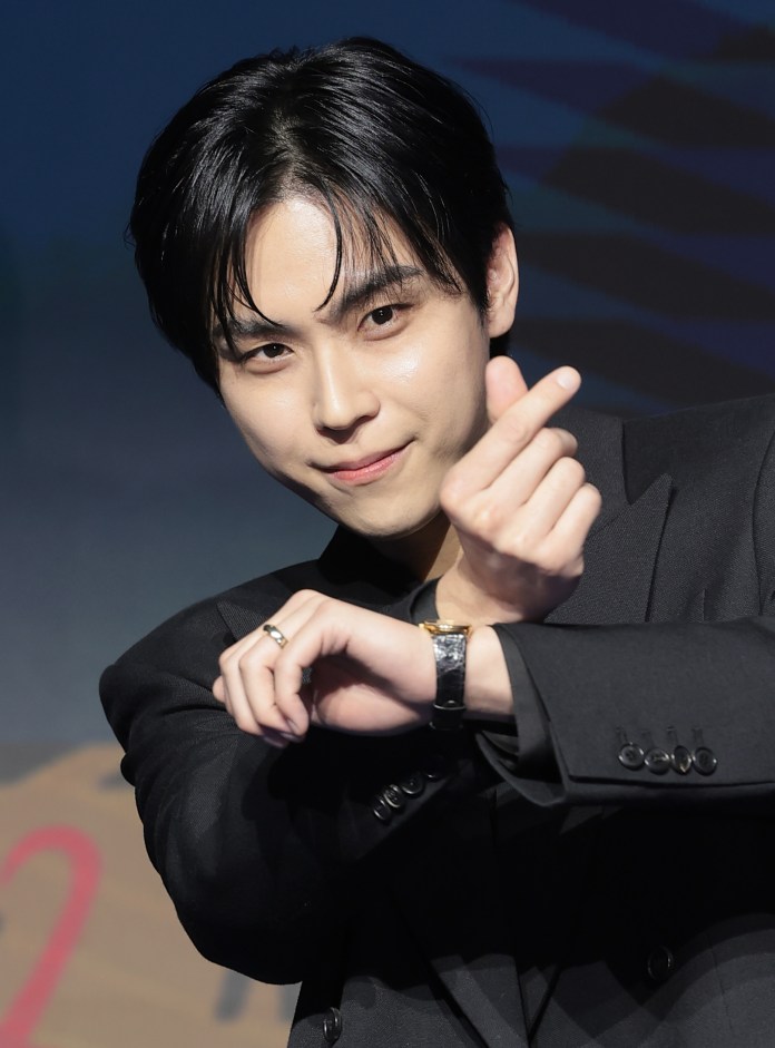 Kim Do-hoon with stylish black hair is posing in a black suit, making a heart shape with his hands and smiling at the camera.