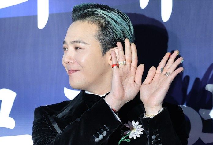 G-Dragon with styled hair and rings on his fingers is smiling and waving with both hands, wearing a black suit with a floral embellishment.