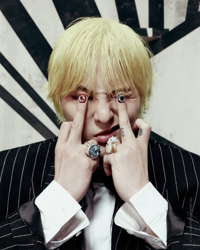 GD with blonde hair makes a playful face, showing two fingers from each hand while wearing multiple rings. They have intricate nail art on their fingers and are dressed in a black pinstriped suit.