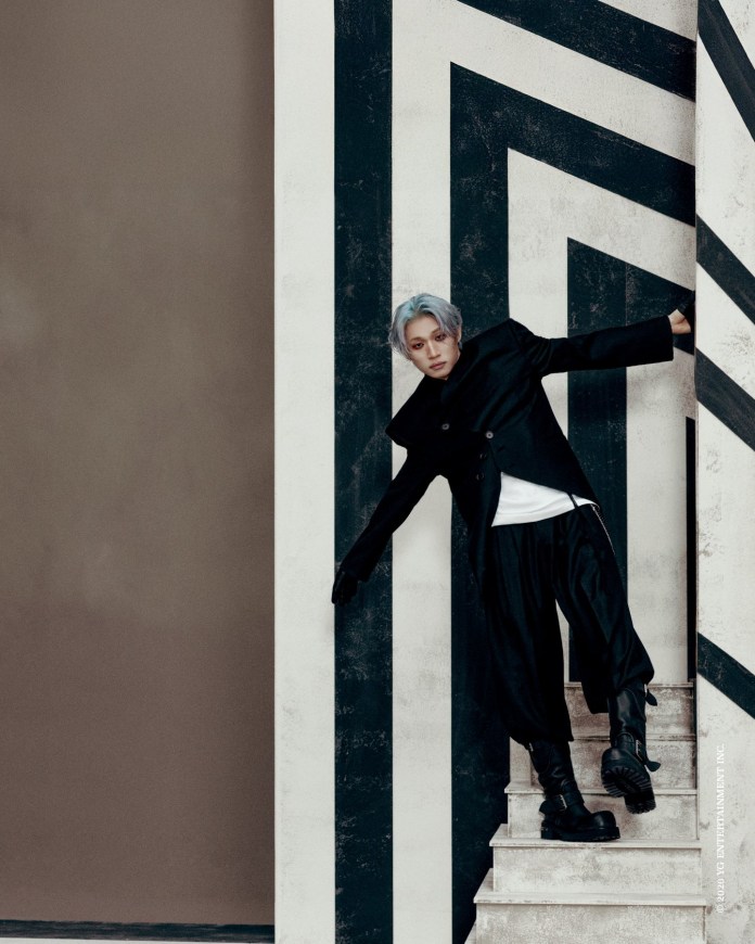 Daesung with silver hair leans against a striped wall while wearing a long black coat, baggy black trousers, and black boots. The background features bold geometric patterns with contrasting colors.