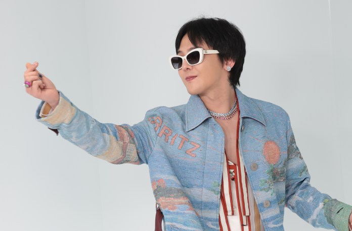 G-Dragon wearing sunglasses and a colorful denim jacket with artistic designs, gestures playfully while posing against a light background.