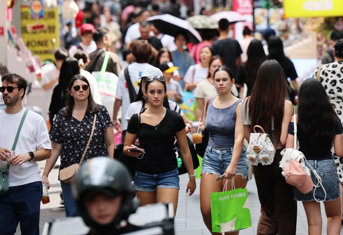 A busy street scene with diverse groups of people walking, some holding drinks and shopping bags, set against a backdrop of shops and signs.