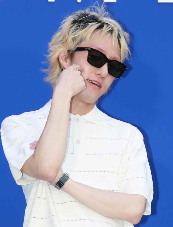 Zion. T with stylish, tousled blonde hair wearing black sunglasses and a white striped polo shirt poses confidently with one hand on his chin and the other arm crossed.