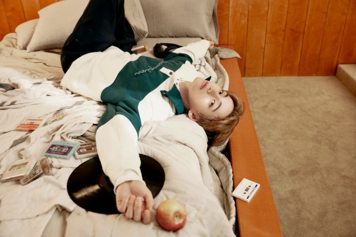 BTS V lying on a bed with a casual outfit, surrounded by cassette tapes, a vinyl record, and an apple, set against a wooden wall.