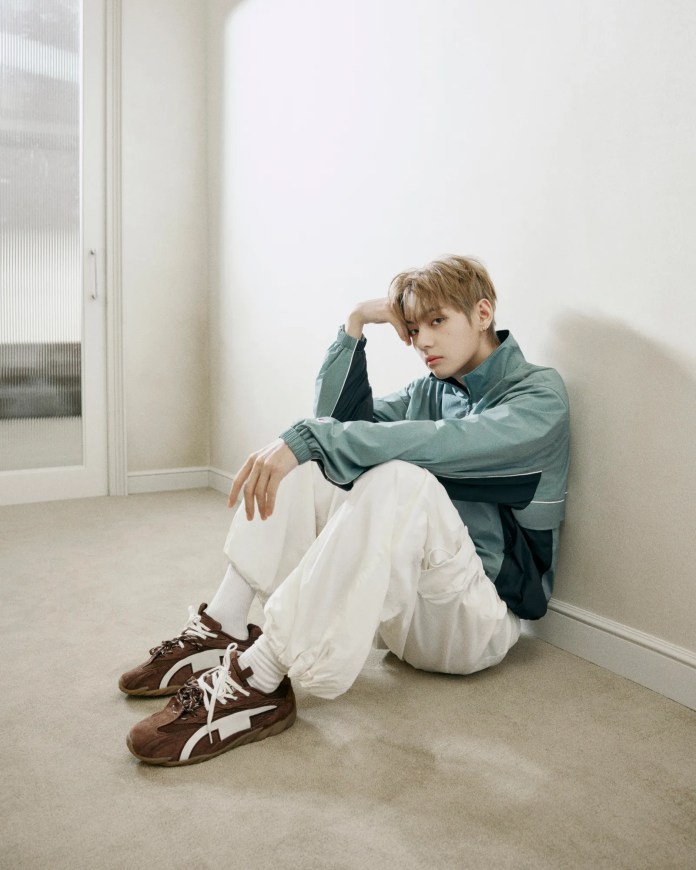 BTS V in a teal jacket and white pants sitting on the floor, looking contemplatively towards the camera, with brown sneakers on his feet and a minimalist background.