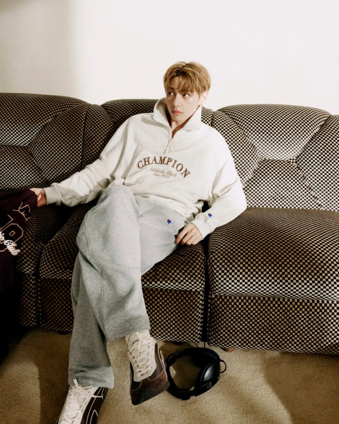BTS V in a casual outfit featuring a white Champion zip-up sweater and gray sweatpants, sitting on a textured brown and black checkered couch, with headphones resting on the floor.