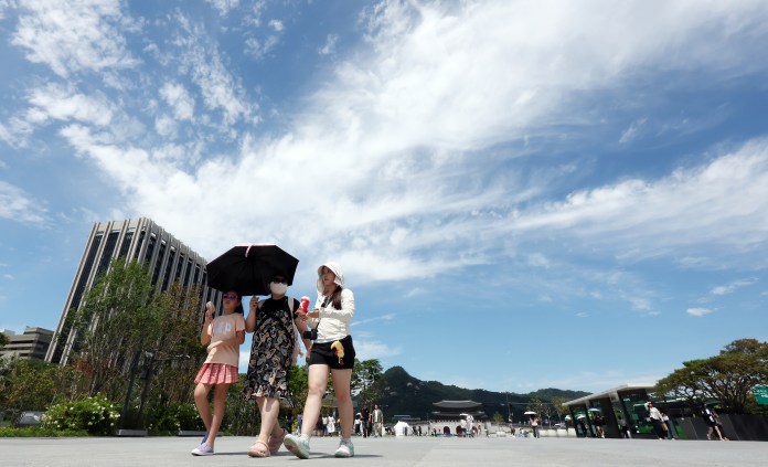 Three individuals walking under a bright sky, one holding an umbrella, with a modern building and greenery in the background.
