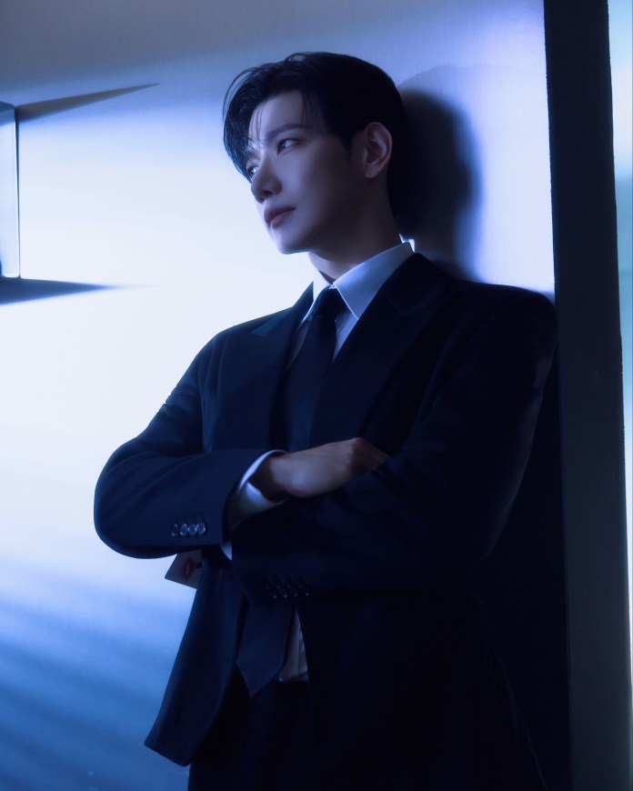 JUN. K in a tailored black suit with arms crossed, leaning against a wall illuminated by soft blue lighting.