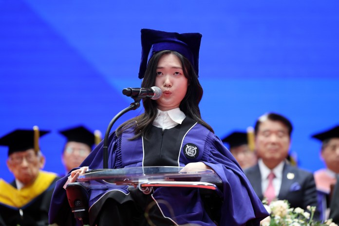 A graduate in a purple graduation gown and cap speaks at a podium during a commencement ceremony, with an audience in the background.