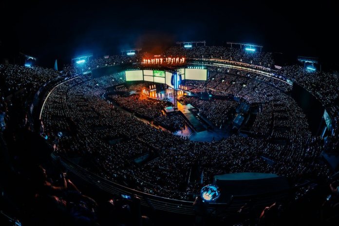 A panoramic view of a large crowd at BTS concert or event, illuminated by stage lights in a stadium setting at night.