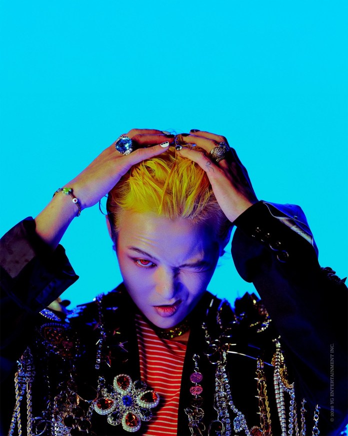 GD with bright yellow hair and a playful expression, wearing a jeweled jacket adorned with chains and a striped shirt. The background is a vibrant blue.