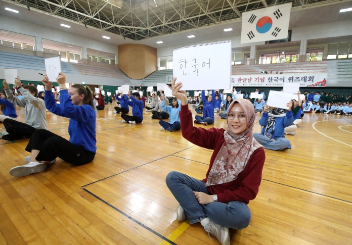A group of students sitting on a gymnasium floor, participating in a language quiz event. Many students are holding signs with Korean text, while one student in the foreground, wearing a hijab and glasses, holds up a sign and smiles.