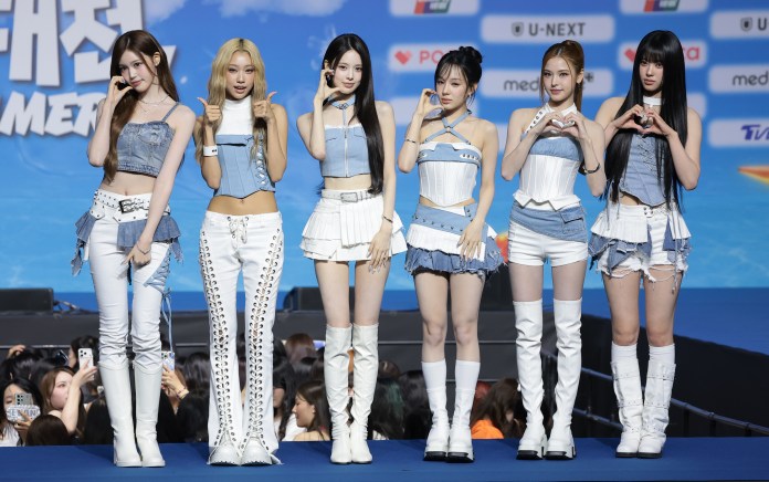 Six women in fashionable outfits posing on stage with a blue background, displaying various hand gestures, including a heart shape.