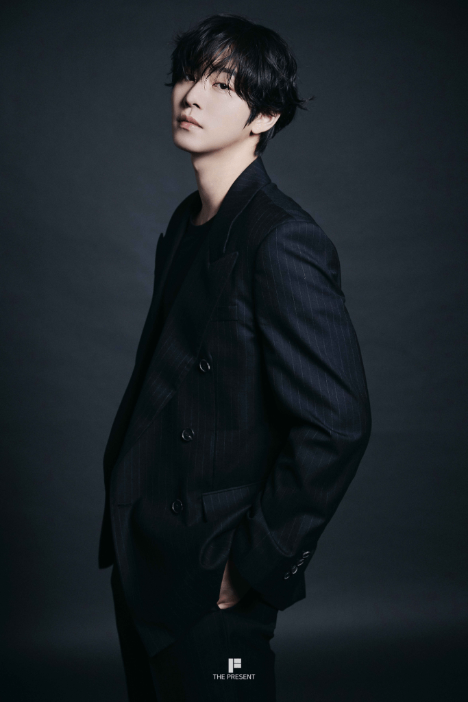 Ahn Hyo-seop in a tailored black suit with pinstripes, standing against a dark background. Their hair is slightly tousled, and they have a contemplative expression.