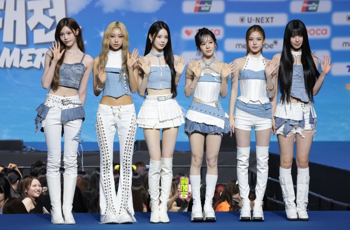 A group of six female performers dressed in coordinated denim and white outfits, posing and waving at a promotional event with a blue background.