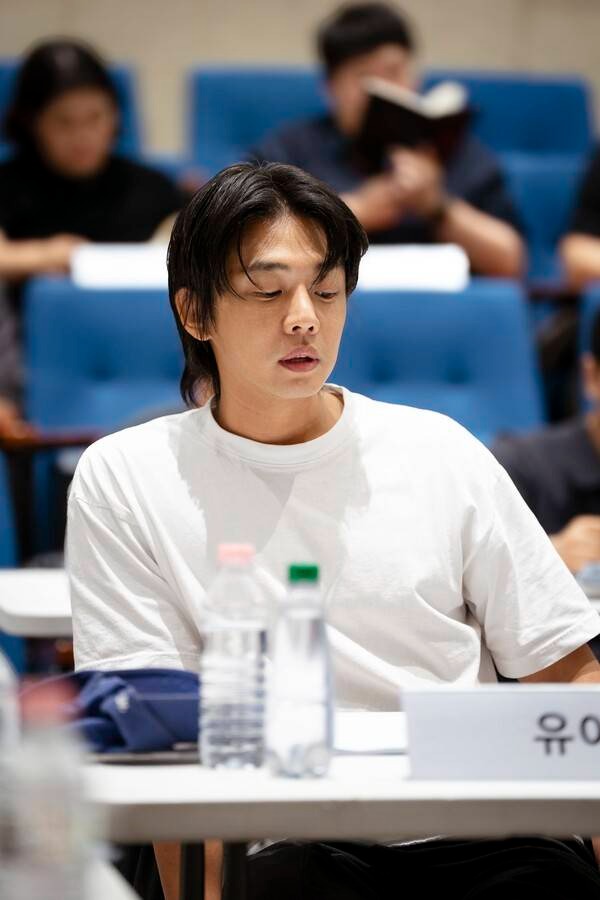 A young man with dark hair wearing a white t-shirt, sitting at a table during a collaborative session. He appears focused and contemplative, surrounded by people in the background who are engaged in reading. Bottled water and a blue bag are visible on the table.