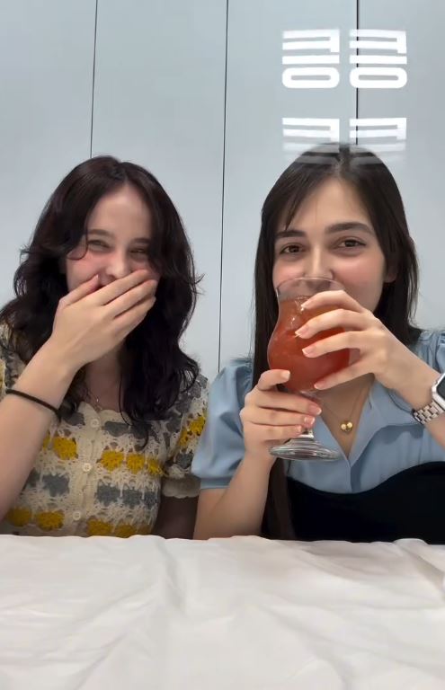 Two women sitting at a table, one laughing and covering her mouth, the other holding a drink with a smile, both enjoying a lighthearted moment.
