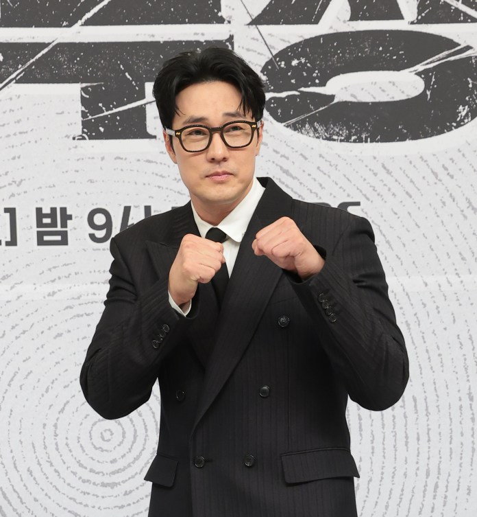A man in a black suit and glasses poses playfully with his fists raised, showcasing a confident expression against a textured background.