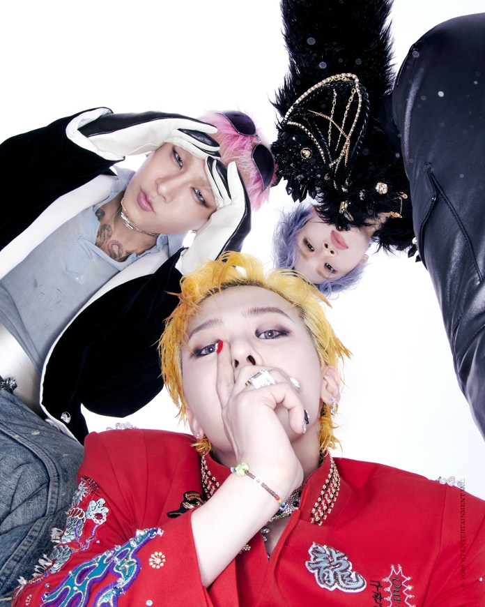 BIGBANG members are seen from a low angle, showcasing diverse hairstyles and colorful fashion. The person in the foreground has bright yellow hair and is making a shushing gesture, while the others have unique hairstyles and outfits, contributing to a striking visual aesthetic.