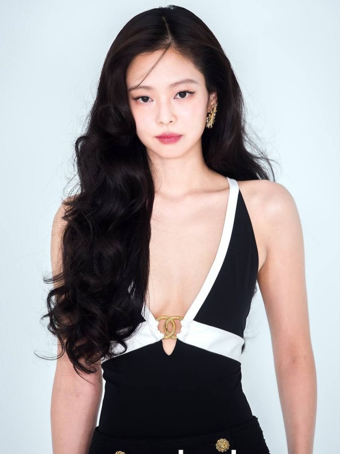 Jennie with long, wavy black hair, wearing a black and white plunging neckline outfit, posing against a light blue background.