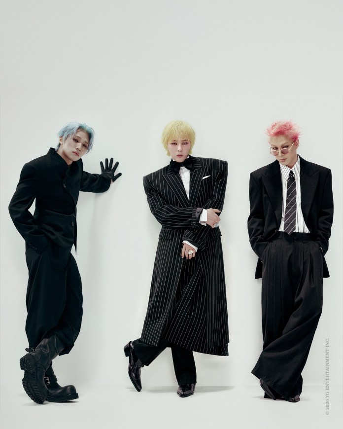 BIGBANG members posing against a plain white background, showcasing unique fashion styles. The person on the left has blue hair and wears a black outfit with bold boots, the center person has yellow hair and a striking pinstripe suit with a bow tie, and the person on the right has pink hair, glasses, and a classic black suit with a striped tie.