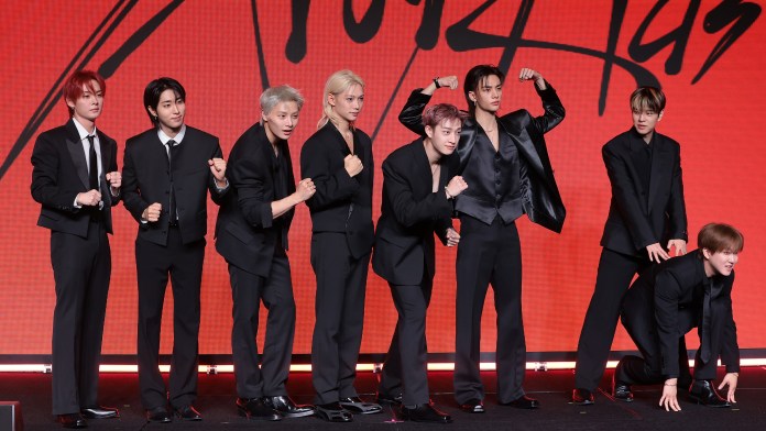 A group of eight men in formal black suits posing together on a stage with a red background.
