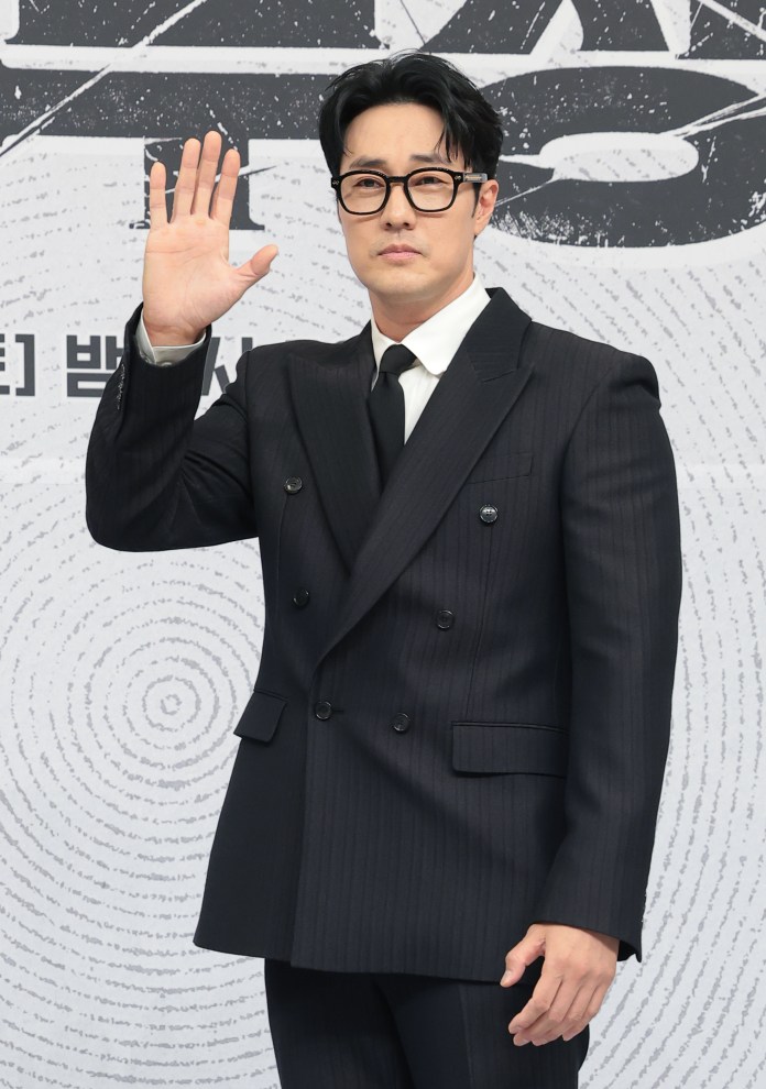 A man in a black double-breasted suit and glasses, waving with his right hand in front of a patterned background.