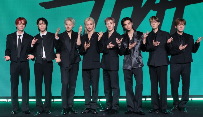 A group of eight men in black suits, standing in a row with various poses, against a green screen background.