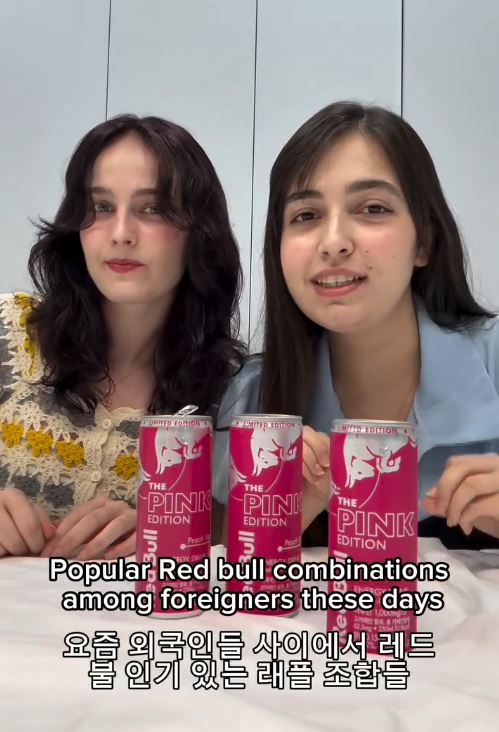 Two young women sitting at a table, smiling and promoting Red Bull Pink Edition drinks, with cans visibly displayed in front of them. The background features neutral-colored walls.