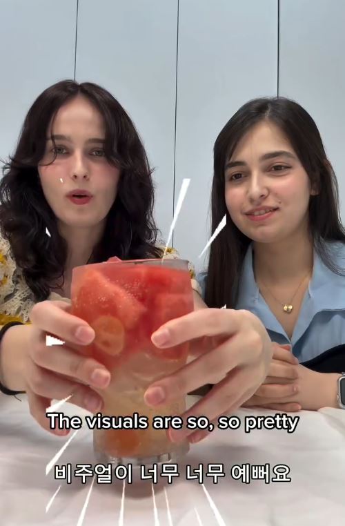Two women sitting at a table holding a colorful drink filled with fruit, expressing excitement about its appearance.